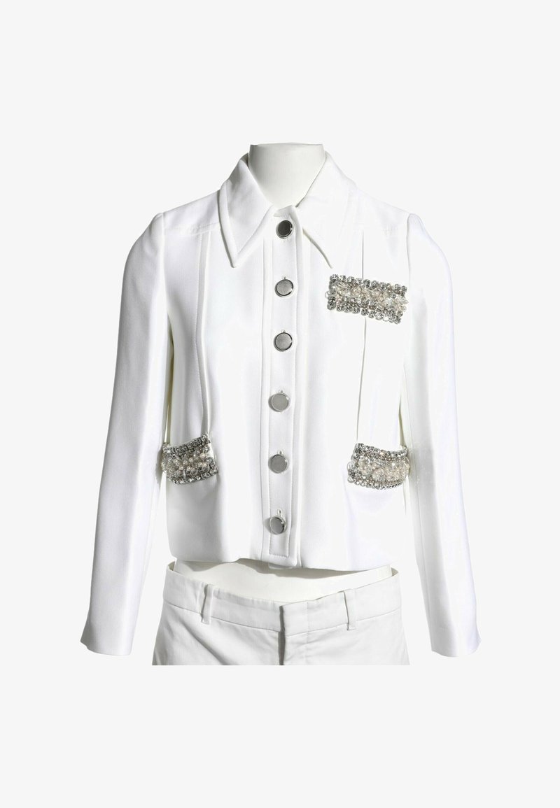 Veste blanche à manches longues avec de grands boutons argentés et trois ornements décoratifs rectangulaires en argent et cristal sur les poches avant et la poitrine.