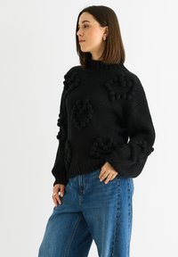 Maglione nero a maglia con motivi a cuore testurizzati e dettagli a pompon, collo alto e maniche lunghe e larghe, abbinato a jeans azzurri chiari.