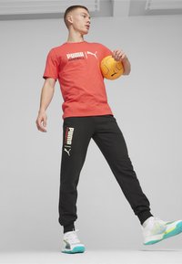 Rode t-shirt met korte mouwen met wit "PUMA HANDBALL" logo, gecombineerd met zwarte joggers met een wit logo. Draagt witte en aqua sport schoenen.