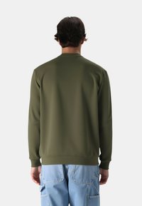 Felpa verde oliva con colletto a girocollo, texture liscia e polsini a coste, abbinata a pantaloni cargo azzurri chiari. La vista posteriore mette in evidenza la semplicità.