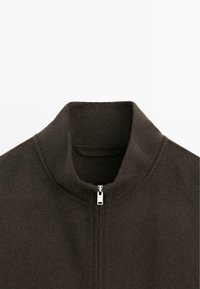 Veste en polaire à col montant de couleur brun foncé, dotée d'une fermeture éclair argentée sur le devant et d'un tissu doux et texturé.