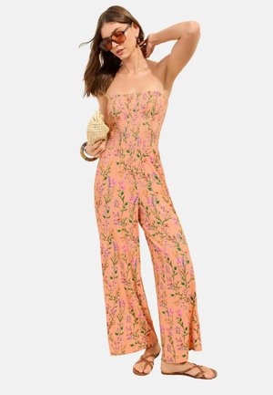 Kvinde iført strapless ferskenfarvet blomsterdragt, brune sandaler, store solbriller, holder flettet clutch, poserer med den ene arm løftet.