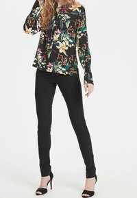 Blommig svart blus med långa ärmar, en knytning i halsen, prydd med vita liljor och färgglada accenter. Kombinerad med åtsittande svarta byxor och klackar.