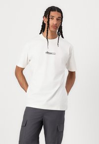 Camiseta de algodón blanca con mangas cortas; presenta el logo de "ellesse" en negro en el pecho; combinada con pantalones cargo grises.