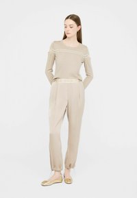 Beige Langarmstricktop mit Schulterauschnitten, kombiniert mit beigen hoch taillierten Hosen mit Zugband. Goldfarbene Ballerinas runden den Look ab.