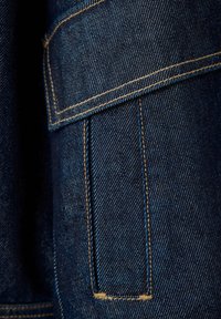 Gros plan d'un tissu en denim bleu foncé avec des surpiqûres jaunes et un détail de rabat de poche.