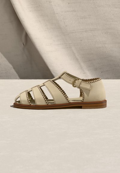 Μπεζ δερμάτινο φλατ σανδάλι με πολλαπλά λουριά, κούμπωμα με αγκράφα, ραμμένες άκρες και καφέ σόλα, σε ανοιχτό φόντο και επιφάνεια.