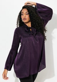 Blusa metallica viola con maniche lunghe e svasate e dettaglio con fiocco al collo. La parte anteriore ha una finitura liscia e testurizzata.