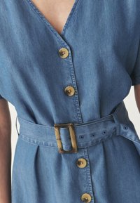 Denimmekko vaaleansinisessä värissä, jossa on V-mallinen kaula-aukko, lyhyet hihat ja vyötäröllä vyö. Sisältää ruskeita nappeja ja teksturoidun pinnan.