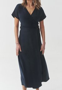 Granatowa sukienka kopertowa wykonana z gładkiego materiału, z krótkimi rękawami, taliowaną linią i ozdobnym guzikiem po bokach.