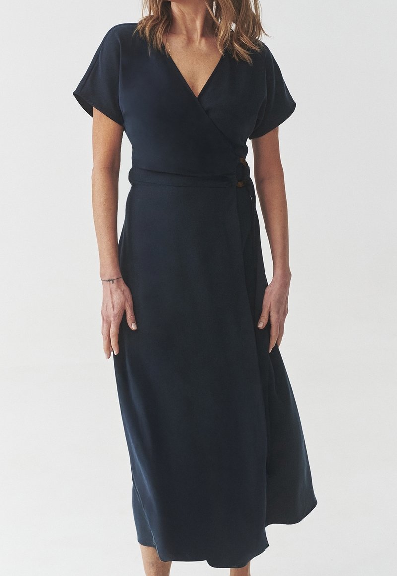 Granatowa sukienka kopertowa wykonana z gładkiego materiału, z krótkimi rękawami, taliowaną linią i ozdobnym guzikiem po bokach.