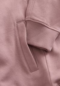 Roosa dressipluusi detail, mis näitab harjatud kangatekstuuri, soonikkoes mansetteid ja küljetaskut puhta õmblustöö viimistlusega.