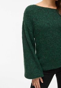 Maglione verde in lana con scollatura ampia e maniche lunghe a campana; tessuto a maglia testurizzato; abbinato a pantaloni neri.