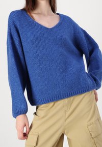 Blå stickad tröja med V-ringning, lös passform och ribbade manschetter, med en texturerad yta och en avslappnad, bekväm design.