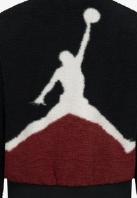Un pull en polaire noir et crème, présentant une silhouette blanche en relief d'une figure sautant, complétée par un accent triangulaire bordeaux en bas.