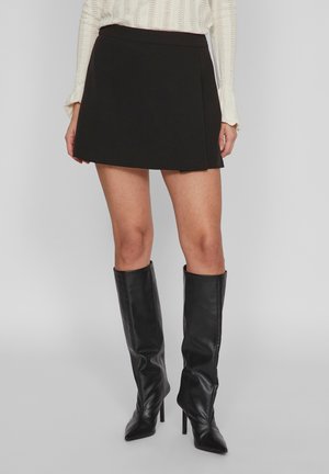 Zwarte mini-jurk met een gladde textuur, met een wikkelontwerp en een aansluitende tailleband, gecombineerd met hoge zwarte leren laarzen.