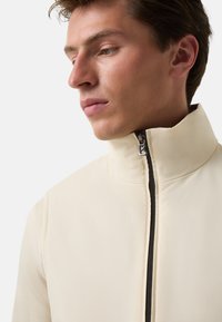Beige Hochkragenjacke aus glattem Stoff, mit einem Frontreißverschluss und dezenten Nähdetails.