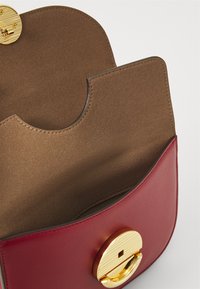 Borsa in pelle rossa e marrone con una patta arrotondata, caratterizzata da una chiusura circolare dorata e una texture interna liscia.
