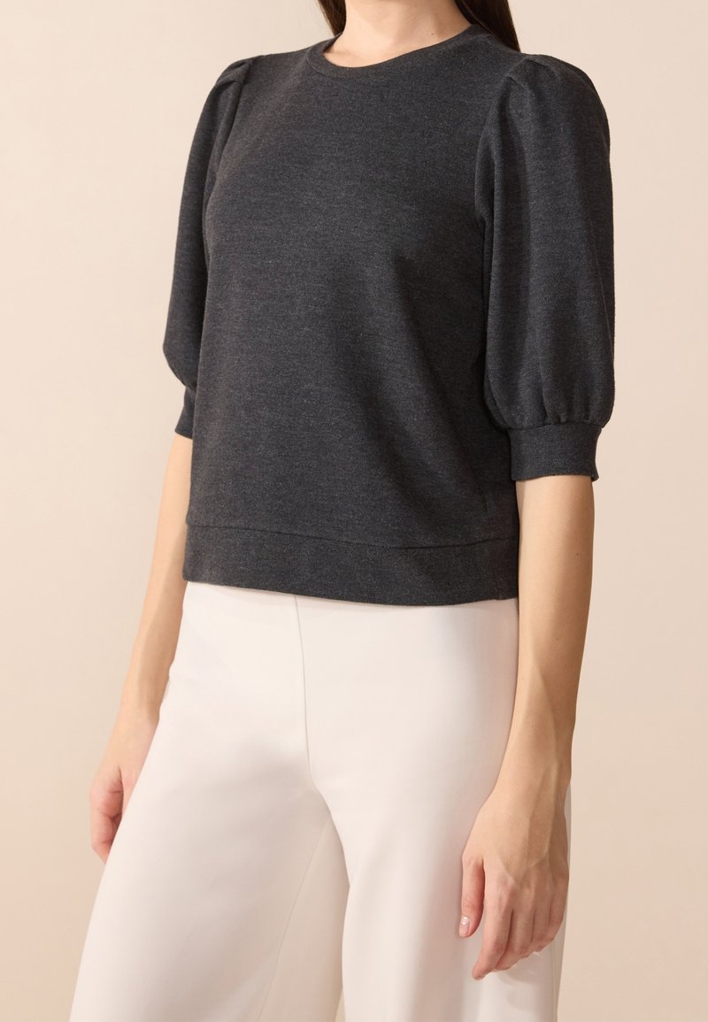 Femme portant un haut gris foncé à manches bouffantes trois-quarts et un pantalon clair taille haute sur fond beige.