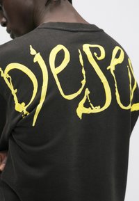 Άνδρας με σκούρο πουκάμισο με μεγάλα κίτρινα, υφή γράμματα με καλλιγραφικό στυλ στην πλάτη, σε φωτεινό φόντο, που έχει γυριστεί ελαφρώς προς τα πίσω.