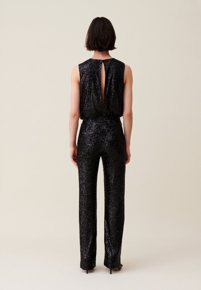 Mono de lentejuelas negras con diseño sin mangas, que presenta un cuello alto y espalda descubierta. Textura suave con una silueta ajustada y piernas anchas.