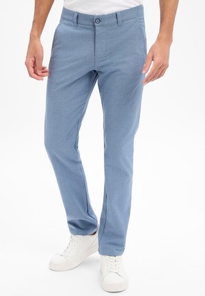 Mężczyzna w jasno-niebieskich, dopasowanych spodniach i białych trampkach, stojący na tle jednolitego białego tła.