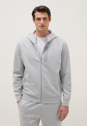 Mann trägt hellgrauen Kapuzenpullover mit Reißverschluss über weißem Hemd und passende graue Jogginghose, steht mit Händen in den Taschen vor einfarbigem Hintergrund.