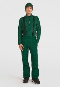 Grön skidoutfit med en zip-up topp och matchande byxor med hängslen. Tyget verkar ha en struktur och byxorna har detalj i form av fickor.