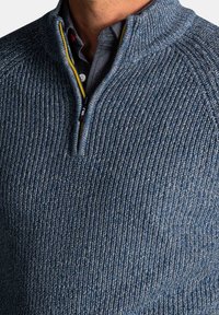 Gebreide trui in gestructureerde blauwe stof met een hoge kraag en halve rits. Heeft rode en gele accenten nabij de rits.