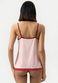 Vrouw met schouderlang donker haar draagt een lichtroze cami met rode kantafwerking en verstelbare bandjes, gezien vanachter.