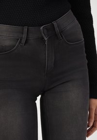 Los pantalones vaqueros de mezclilla negra presentan un diseño ajustado, estilo de cinco bolsillos y un cierre frontal con botón. La tela tiene una textura suave.