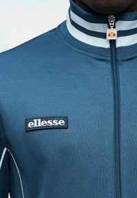 Μπλε αθλητικό τζάκετ με φερμουάρ και υψηλό γιακά, με λευκές ρίγες και μαύρο κεντημένο λογότυπο «ellesse» στο στήθος. Ομαλή υφή.