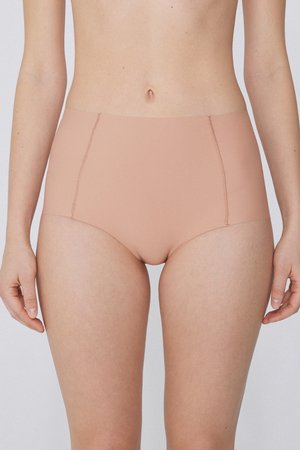 Žena v bezšvém tělovém spodním prádle s vysokým pasem, stojící s rameny uvolněnými proti jednobarevnému pozadí.