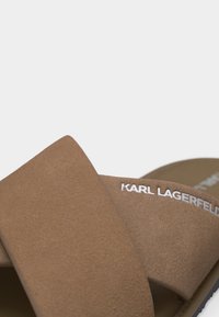 Καφέ σουέντ λουριά σανδαλιού που σχηματίζουν σταυρό με την επιγραφή "KARL LAGERFELD" σε λευκό σε ένα λουρί, κοντινή λήψη.