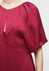 Κοντινό πλάνο ενός ατόμου που φοράει ένα βαθύ κόκκινο σατέν φόρεμα με κοντά puffed μανίκια και λεπτομέρεια κόμπου μπροστά κοντά στη γραμμή του λαιμού.