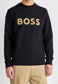 Hombre con sudadera negra con gran texto dorado "BOSS" y pantalones beige, de pie frente a un fondo claro y liso.