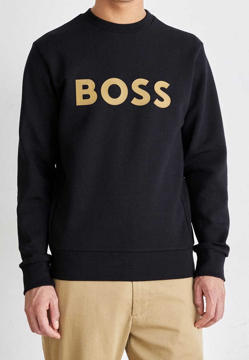 Hombre con sudadera negra con gran texto dorado "BOSS" y pantalones beige, de pie frente a un fondo claro y liso.