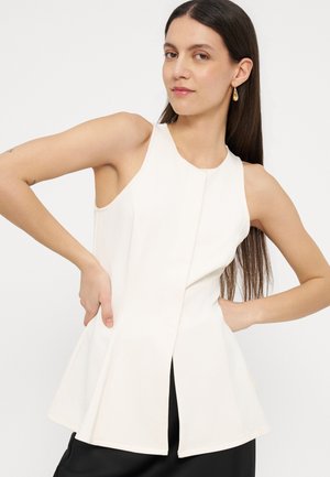 Žena s dugom tamnom kosom, odjevena u bijelu brezrukavu peplum majicu i zlatne naušnice na kapljicu, pozira s rukama na bokovima na bijeloj pozadini.