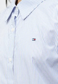 Biela a modrá pruhovaná košeľa na gombíky vyrobená z bavlny, s klasickým golierom a malou nášivkou v červenej, bielej a modrej farbe.