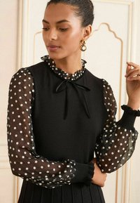 Black dress with a high ruffled neckline, long sheer sleeves adorned with white floral patterns, and a tied satin ribbon at the collar.