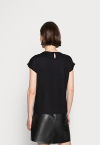 Kvinna med kort mörkt hår iklädd en svart kortärmad topp med nyckelhålsdetalj och svart läderkjol, vänd bort från kameran mot en enfärgad bakgrund.