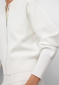 Veste blanche zippée en tissu texturé. Présente des poignets et un ourlet côtelés. Design minimaliste avec une silhouette ajustée et sans motifs visibles.
