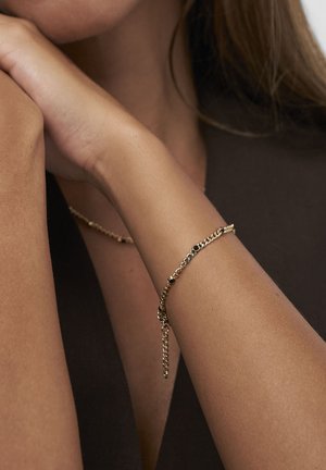 Goldene Kettenarmband mit schwarzen Steinen, das an einem Frauenhandgelenk vor einem dunkelbraunen ärmellosen Oberteil getragen wird.