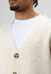 Beige gebreid vest met een V-hals, voorzien van twee bruine knopen en geribbelde randen. Draagt over een wit T-shirt met ronde hals.