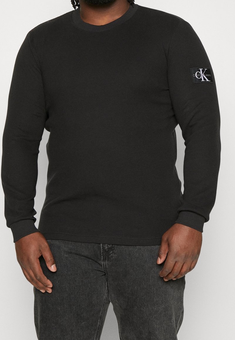 Chemise noire à manches longues en texture côtelée, avec un col rond et un patch logo sur la manche gauche. Associée à un jean foncé.