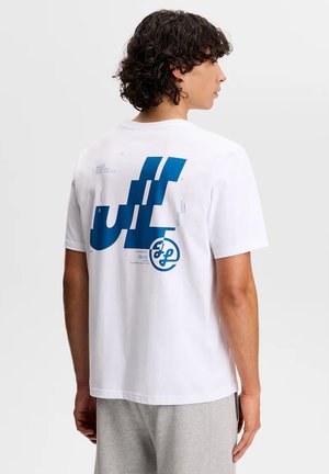 Persona con cabello rizado que lleva una camiseta blanca con un gran gráfico abstracto azul y texto en la parte posterior, combinada con pantalones gris claro.