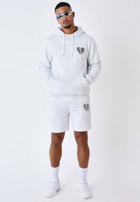 Sudadera gris y pantalones cortos a juego de algodón, con un gráfico de corazón roto. Modelo con zapatillas blancas y gafas de sol.
