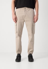Beige cargobroek met een elastische tailleband, twee zijvakken en taps toelopende pijpen. Gecombineerd met zwarte sneakers. Glad materiaal.