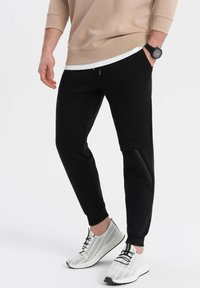 Schwarze Jogginghose mit seitlichen Reißverschlüssen, gerippter Textur, tapered Fit, kombiniert mit einem beigen Sweatshirt und weißen Mesh-Sneakern.