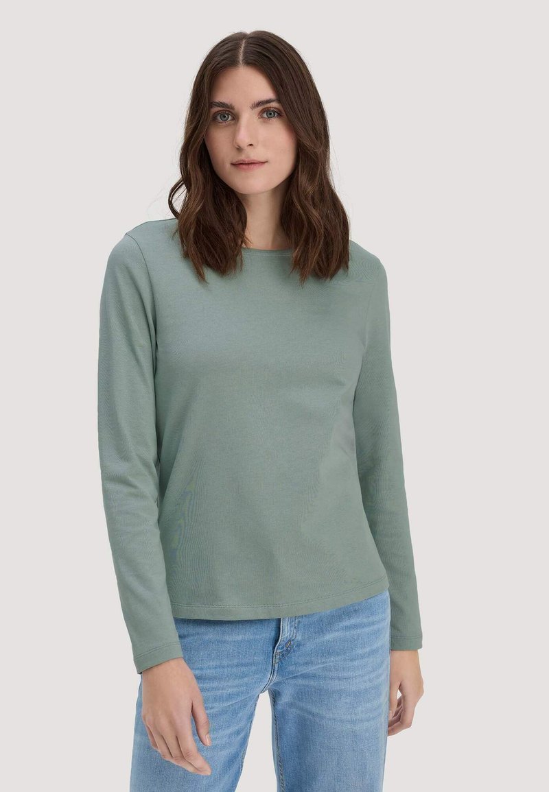 Jeune femme aux cheveux bruns ondulés portant une chemise verte sauge à manches longues et un jean bleu clair, debout devant un fond uni.